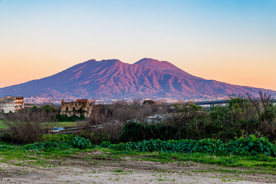 A breathtaking view of Mount Vesuvius during sunset, with soft orange and pink hues illuminating the majestic volcano. The foreground features rural ruins and greenery, creating a serene and timeless 