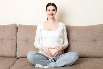 Fototapeta premium photo of cute caucasian future mom lying in light living room on sofa and holding hands on tummy