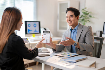 A man and a woman are sitting at a desk with a calculator in front of the man