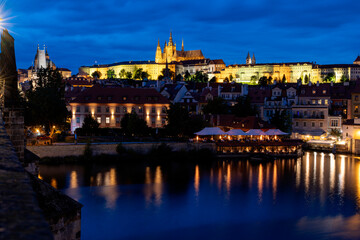 A picturesque view of Prague Castle and surrounding historical architecture, with reflections on the Vltava River under a cloudy sky