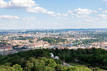 A wide-angle view of Prague’s iconic red rooftops, historic churches, and lush green surroundings. The cityscape combines traditional architecture with glimpses of modern structures in the background