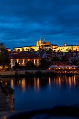 A picturesque view of Prague Castle and surrounding historical architecture, with reflections on the Vltava River under a cloudy sky