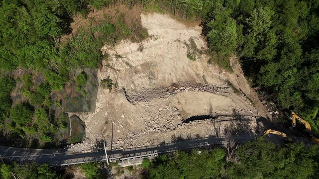 Aerial slow pan shows a large landslide where the terrain collapsed, disrupting a road along the forest. Earth-moving machinery is visible near the affected area near Vernasca, PC, Italy