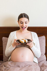 Hungry pregnant woman is eating tasty desserts staying in bed at home. Enjoying sweet food during pregnancy concept