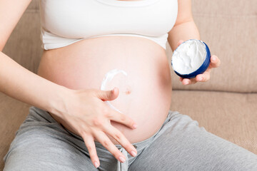 a pregnant girl sits at home on the bed and smears heart an anti-stretch mark cream on her stomach. Pregnancy, motherhood, preparation and expectation concept