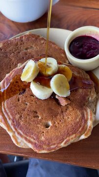 Maple syrup is poured on pancake with banana for breakfast in cafe or restaurant. Fancy bistro shop with delicious menu filmed from phone pov of foodie person or influencer eating out in city downtown