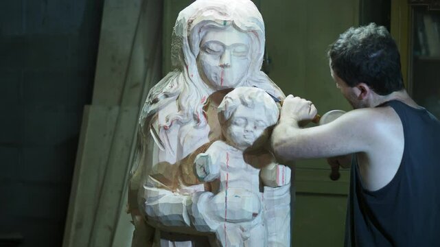 A wood sculptor hand-carves a large statue depicting the Madonna and Child using chisel and mallet. Red guide lines mark the contours as he shapes the religious figure in his workshop, slow motion.
