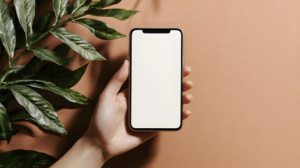 A hand holds a smartphone with a blank screen against a beige background next to green leaves.