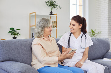 Fototapeta premium Young cheerful female caregiver teaching senior person to handle with mobile phone at home. Friendly nurse showing elderly woman how to use smartphone. Nursing home service and patient support.