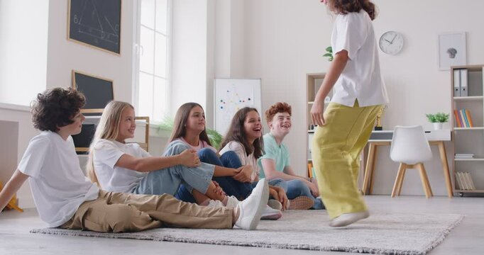 Group of happy school children having fun playing charades indoors during recess playtime. Cheerful joyful kids sit on floor inside classroom, looking at funny girl, guessing who or what she is miming