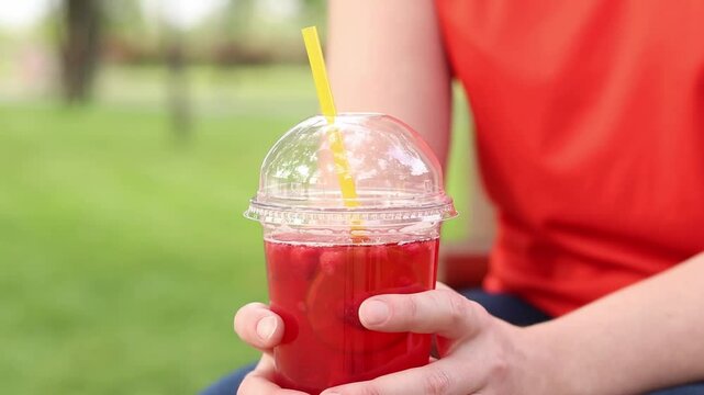 Close-up of female hands with lemonade in a disposable cup. Summer drink. Girl in a bright T-shirt sits in the park. Summer and recreation concept. Pink lemonade with raspberry and lime
