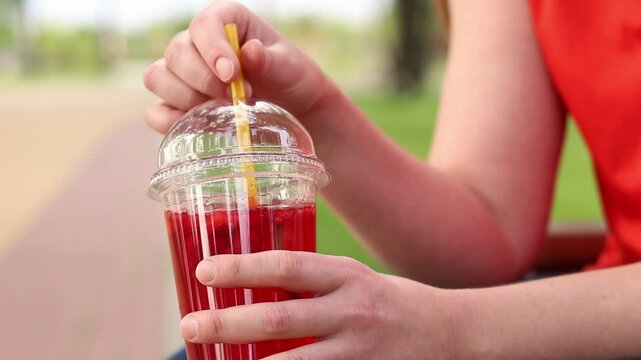 Close-up of female hands with lemonade in a disposable cup. Summer drink. Girl in a bright T-shirt sits in the park. Summer and recreation concept. Pink lemonade with raspberry and lime