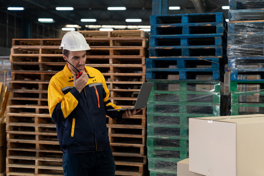 Adult male warehouse supervisor with light skin wearing white helmet and yellow jacket, using walkie-talkie and holding laptop, standing in front of stacked pallets and boxes in storage facility.