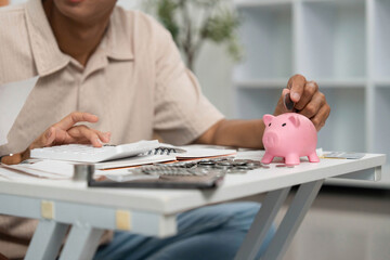 Financial Management and Savings. Young man depositing coins into a piggy bank at home.