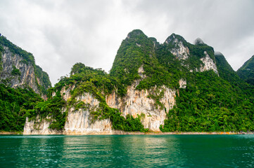 View of Khao Sok Lake in Krabi province, Thailand, with towering limestone cliffs covered in tropical rainforest rising above the calm surface of the lake.