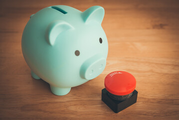 Blue piggy bank looking at red stop emergency button on wooden table background. Home finance or...