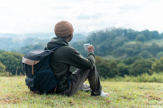 An Indonesian man sits on the grass with a backpack, sipping coffee while enjoying a serene mountain landscape. Captures peaceful travel moments, solitude, and connection with nature.