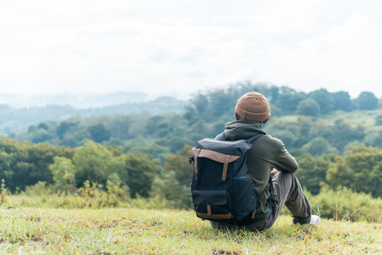 An Indonesian man sits on the grass with a backpack, sipping coffee while enjoying a serene mountain landscape. Captures peaceful travel moments, solitude, and connection with nature.