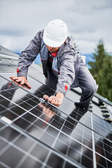 Man roofer mounting photovoltaic solar panels on roof of house. Engineer in helmet installing solar module system with help of hex key. Concept of alternative, renewable energy.