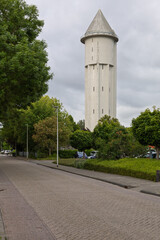 The iconic water tower of Meerkerk, Netherlands, stands tall under a cloudy sky, a distinctive landmark amidst green trees and a quiet street. Built in 1936.