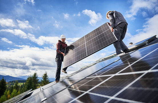 Roofers building solar panel system on roof of house. Men workers in helmets carrying photovoltaic solar module outdoors. Concept of alternative and renewable energy.