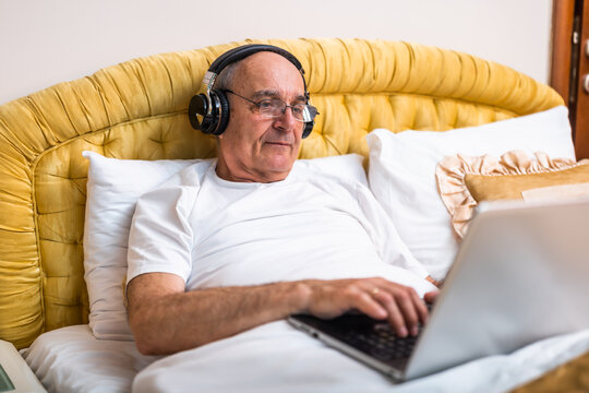 
Relaxed senior man in pajamas with headphones using laptop while enjoying resting in bed at his home.