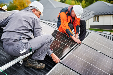 Roofers building photovoltaic solar module station on roof of house. Men electricians in helmets installing solar panel system outdoors. Concept of alternative and renewable energy.