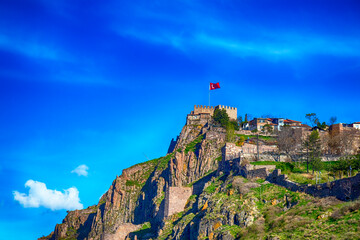Historic Ankara Castle perched on rocky cliffs under a vibrant blue sky with Turkish flag waving...