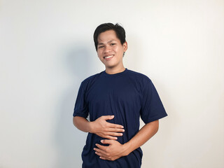 A cheerful young Asian man wearing a blue shirt smiles while holding his stomach, standing against...