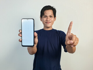 Young Asian Man Smiling While Pointing at Smartphone with Blank Screen, Casually Dressed in Blue...