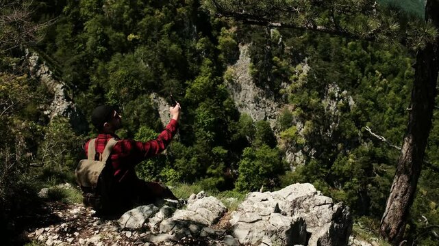 Man sits at Banjska stena viewpoint overlooking Drina river in Tara National Park, Serbia, holding phone and enjoying panoramic spring mountain scenery. Solo travel concept. Outdoor blogger