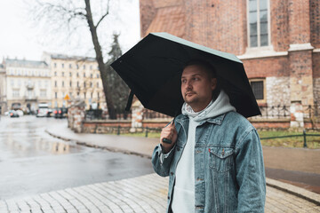 Man in a denim jacket and hoodie walks through a rainy European street, holding a large black...