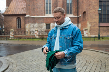 Obraz premium Man in a denim jacket and hoodie is seen folding a green umbrella on a cobblestone street, with a historic brick building in the background. The wet pavement hints at recent rain, creating a peaceful