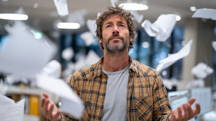 A stressed professional stands before a printer, hands on head, surrounded by flying papers. Cool lighting and blurred office background highlight the chaos and pressure of workplace overload.
