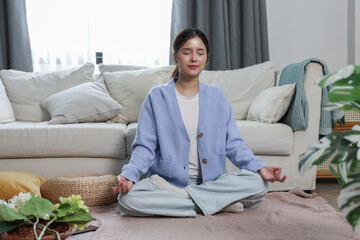 Young woman meditating peacefully at home, sitting cross legged on carpet, practicing mindfulness and relaxation in cozy living room environment