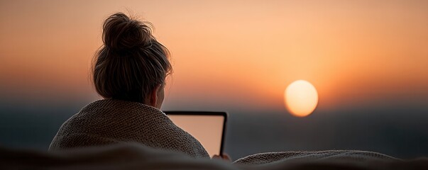 A person wrapped in a blanket works on a laptop while watching the sunset in a serene outdoor setting.