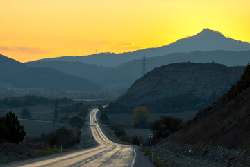 A long winding road stretches through mountainous terrain during sunset, with glowing yellow sky and hills in the background, capturing the beauty of nature and open road travel.

