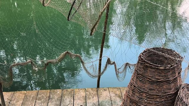 A wooden pier with wicker baskets for fish on it and antique fishing nets hanging on the pier against the backdrop of the lake.