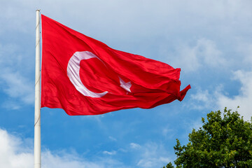 Turkish flag fluttering vividly against a partly cloudy blue sky, symbolizing Turkey's national pride and heritage, surrounded by green trees in an outdoor setting.

