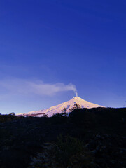 Large volcano covered in white snow, among the Araucaria forests, constantly releasing smoke from...