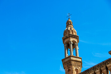 A detailed bell tower in Midyat, Mardin stands tall against a vibrant blue sky. The intricate stonework and cross highlight the rich Christian heritage and architectural beauty of this historic region