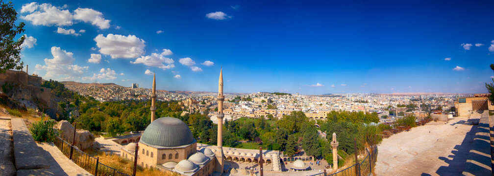 a panoramic view of şanlıurfa city taken from şanlıurfa castle, featuring mosque minarets, lush greenery, and dense city buildings under a bright blue sky with scattered clouds