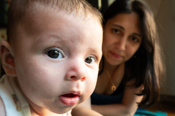 Baby peers into camera while mother’s soft smile appears in background; warm indoor light highlights expressive eyes and tender family connection.