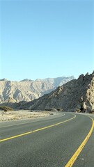 Road curves beneath towering cliffs as shadows ripple across rugged terrain, cliffs rise sharply from ancient layers, forming deep rhythm carved into raw geological landscape.