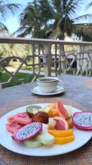 Fresh tropical fruit platter beside steaming coffee mug, resting on resort balcony railing with verdant palm landscape stretching below, capturing peaceful morning moment