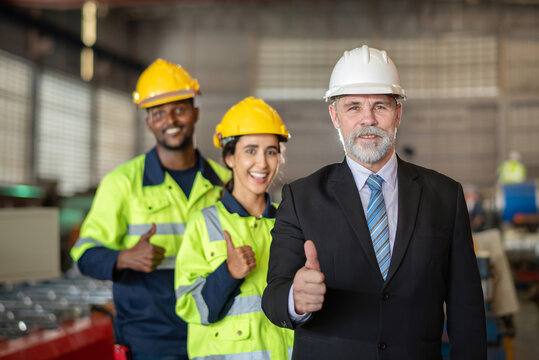 mature older boss engineer with beard mustache on face wear helmet standing smile and hand show good thumb at construction factory site with worker leader team for happy working in factory site.