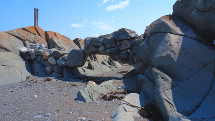 An arch made of an agglomeration of old rocks connecting two geological formations on the shore of a sandy beach under a blue sky.