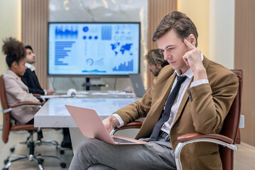 A man in a brown suit jacket and dark colored dress shirt and tie is sitting in an office space...