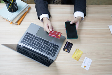 A man is using a laptop and a cell phone to pay for something. He is holding a credit card and a debit card
