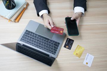 A man is using a laptop and a cell phone to pay for something. He is holding a credit card and a debit card
